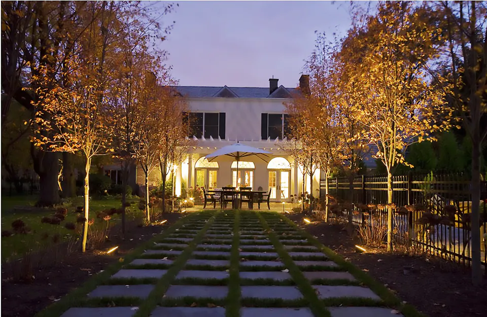 Back of a house at night with uplit trees, a lit patio with an umbrella, and a stepping-stone walk through lawn